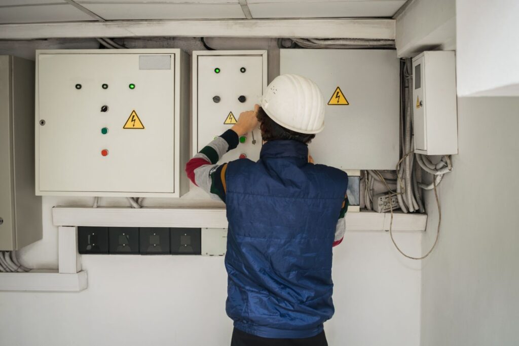 A man in a hard hat and vest inspects electrical equipment in a basement construction site