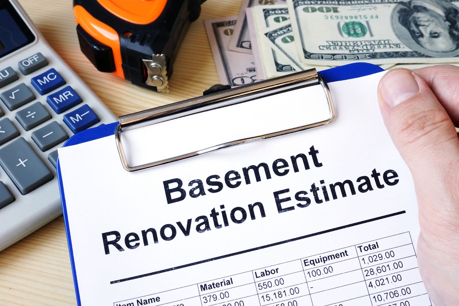 Hand holding a clipboard with a basement renovation estimate, showing costs for materials, labor, and equipment, next to a calculator, tape measure, and cash.