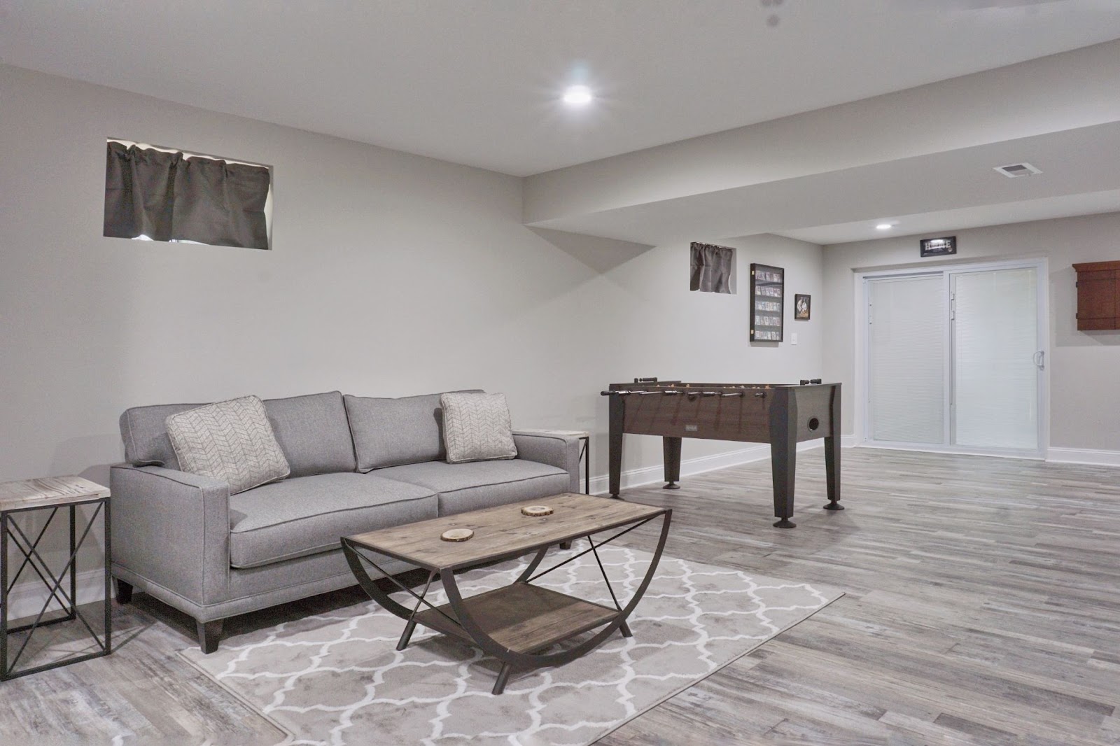 Modern basement room with a gray couch, coffee table, area rug, and foosball table on gray wood flooring.