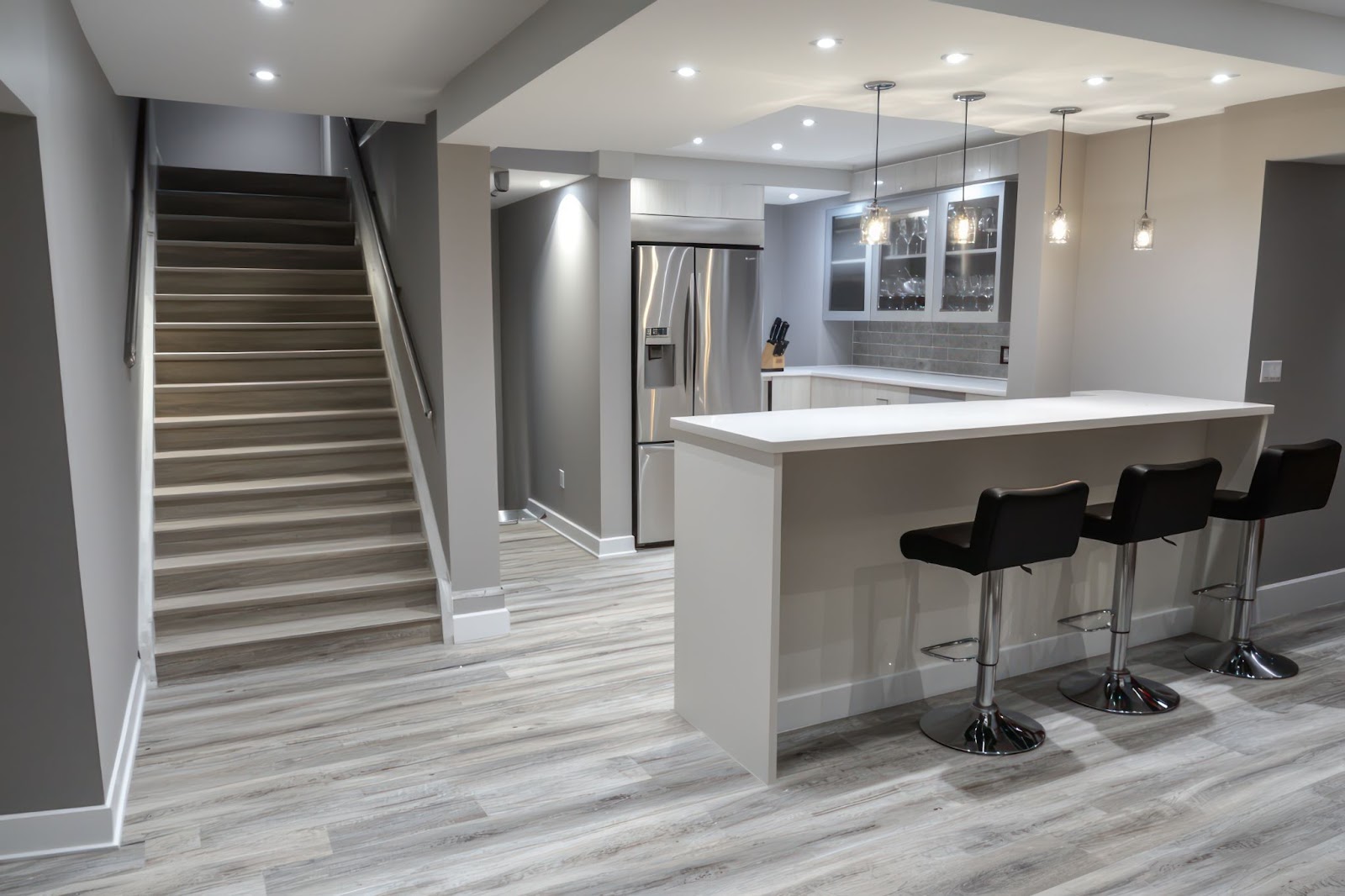 Modern basement with a kitchen bar, black stools, and wooden stairs.