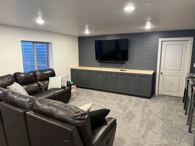 Cozy basement media room with dark gray shiplap accent wall, mounted TV, long gray built-in cabinet with wood top, brown leather sectional sofa, light gray carpet, and recessed lighting.