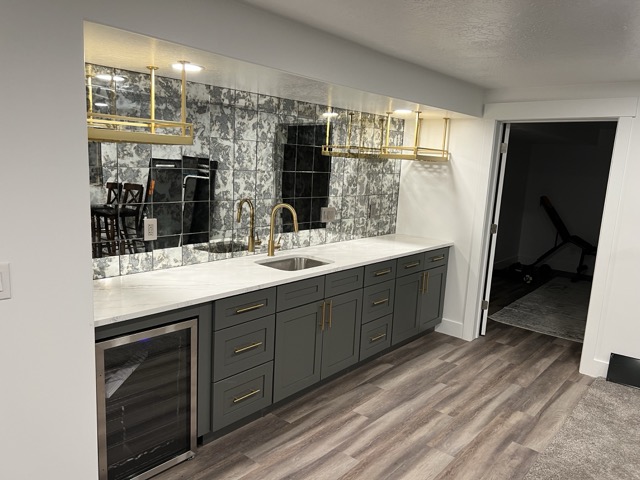 Modern basement bar with dark green cabinets, gold handles, white marble counter, antique mirror tile backsplash, gold hanging glass racks, under-counter wine fridge, and wood-look flooring.