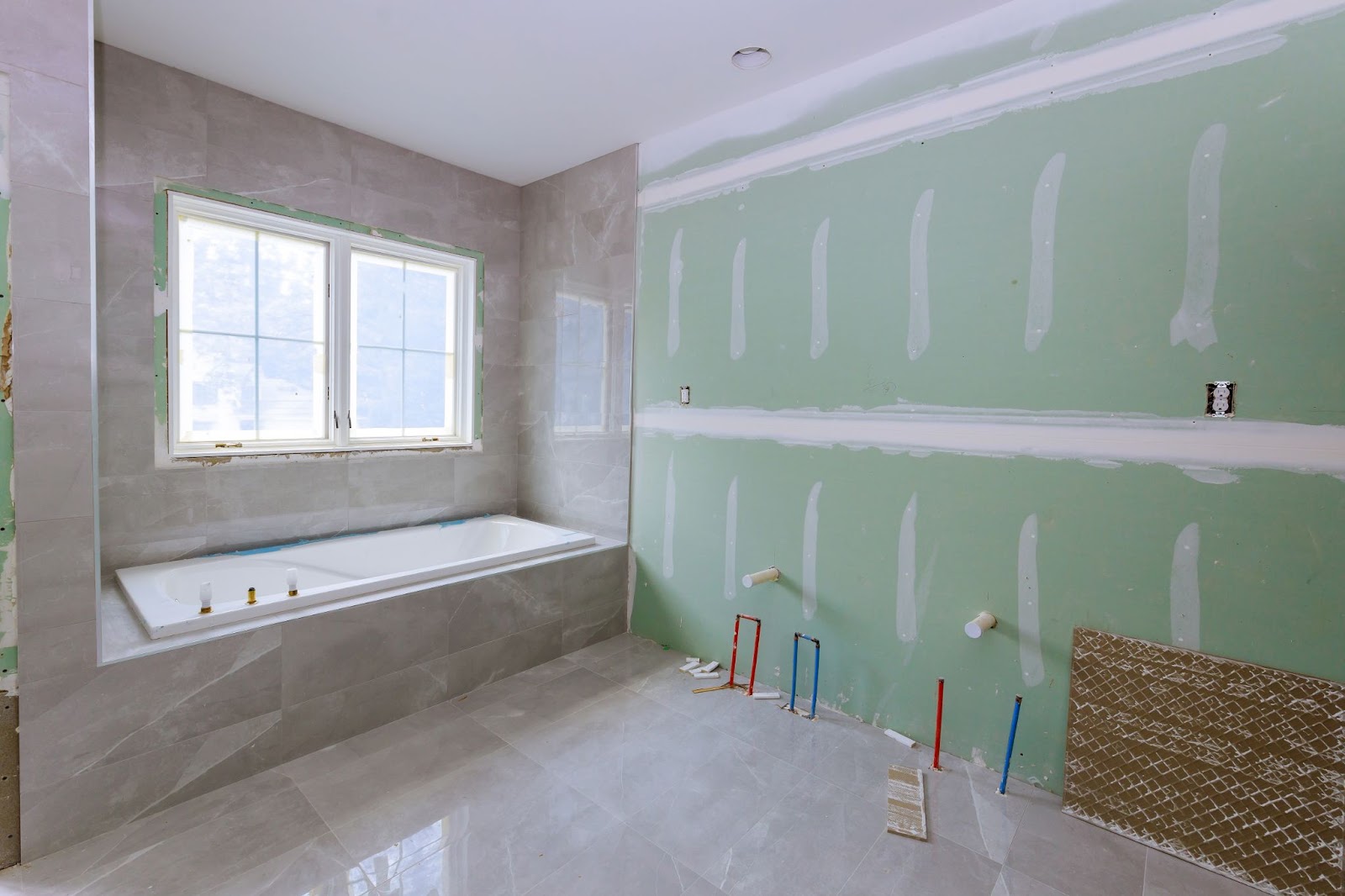 A bathroom under renovation featuring a white bathtub surrounded by large grey floor and wall tiles, with one wall of green drywall showing exposed plumbing lines.