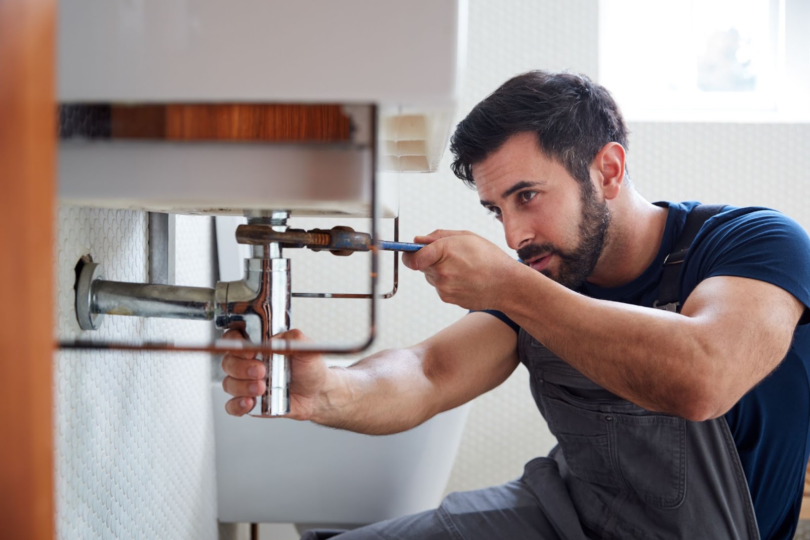 A plumber in grey overalls uses a wrench to tighten a chrome pipe beneath a white bathroom sink.
