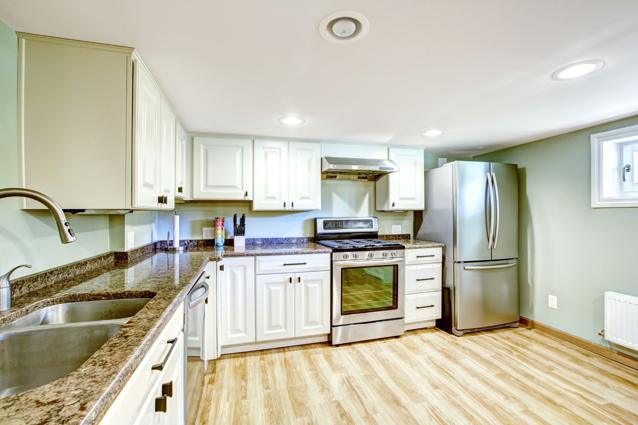 A basement kitchen featuring white cabinetry, granite countertops, stainless steel appliances, and light wood flooring.