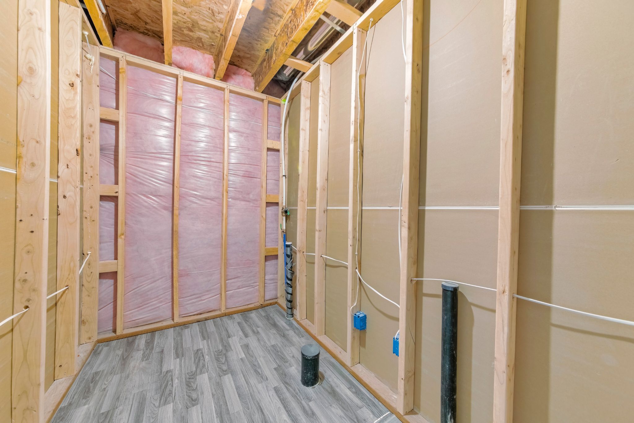 An unfinished bathroom or room under construction, showing wood wall framing, pink fiberglass insulation, exposed electrical wiring, and rough-in plumbing drains over a grey wood-look floor.